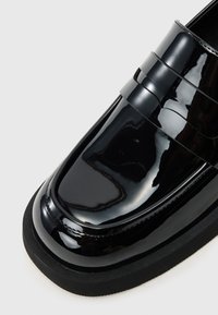 Mocassin en cuir verni noir avec une surface lisse et brillante, présentant un détail de lanière surélevé et une semelle en caoutchouc robuste.