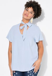 Blusa azzurra chiara con scollo a V, maniche a volantino e dettaglio della fascia attorno al collo. Realizzata in un tessuto liscio e morbido.