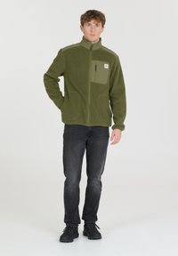 Grüner Fleece-Jacke mit hohem Kragen, Reißverschluss vorne, Seitentaschen und einer Brusttasche. Kombiniert mit schwarzen Jeans und Sneakern.