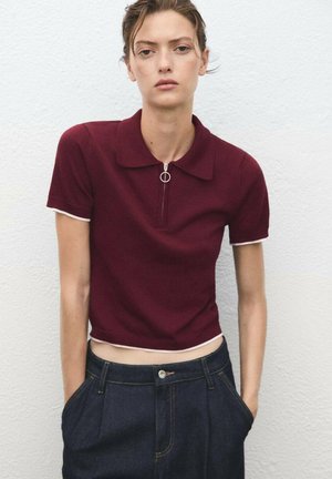 Burgundská polo tričko s krátkým zipem a stříbrným detailním kroužkem, s bílým lemováním na rukávech a mírně zkráceným střihem. V kombinaci s tmavými denimovými kalhotami.