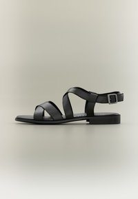 Černé kožené sandály s plochou podrážkou, charakterizované designem překřížených pásků a nastavitelnou přezkou u kotníku pro bezpečné upevnění.