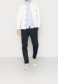 Witte katoenen jas met bruine knopen over een lichtblauw overhemd, gecombineerd met donkerblauwe broek en beige sneakers, staand tegen een effen achtergrond.