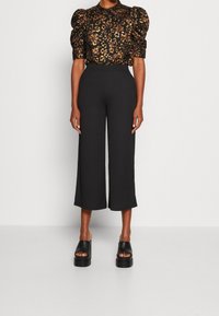 Blouse noire à motifs floraux avec des manches bouffantes, associée à un pantalon noir large taille haute et des sandales plateformes noires épaisses.