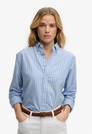 Mujer rubia con las manos en los bolsillos, que lleva una camisa de botones a rayas azul y blanca, metida dentro de unos pantalones blancos con un cinturón trenzado marrón.