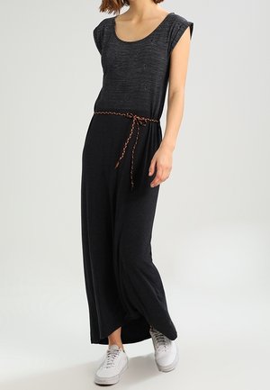 Mujer con un vestido maxi negro sin mangas con un cinturón trenzado y zapatillas blancas, de pie contra un fondo claro y sencillo.
