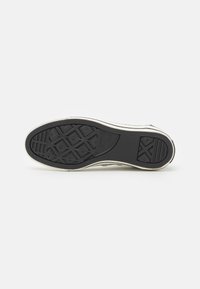 Semelle de chaussure de sport en caoutchouc noir avec motif de grip texturé, bordée d'un bord blanc. Elle présente un motif en losange et plusieurs rainures pour une meilleure traction.