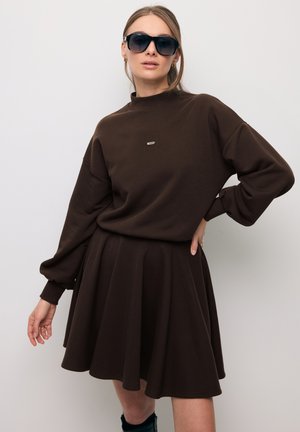 Vrouw draagt een donkerbruine jurk met lange mouwen, uitlopende rok en oversized mouwen, zwarte zonnebril, staand met één hand op haar heup.