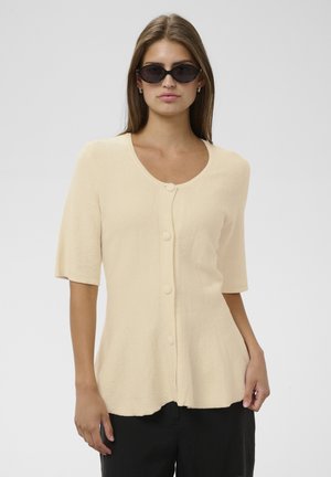 Femme portant un cardigan beige à manches courtes boutonné et un pantalon noir, avec de longs cheveux et des lunettes de soleil ovales noires, debout devant un fond uni.