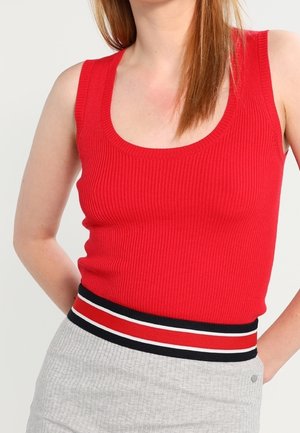 Vrouw draagt een mouwloze rode geribde tanktop, gestopt in een lichtgrijze rok met een tailleband met zwarte, rode en witte strepen.