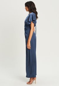 Langes navy-blaues Satinkleid mit kurzen Puffärmeln, gerafftem Detail an der Taille und einem Schlitz an der Seite, kombiniert mit nudefarbenen Schnürsandalen.