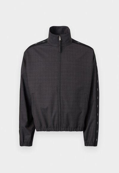 Jachetă gri închis cu fermoar, cu guler înalt, manșete și tiv elastice, și dungi negre cu detalii albe de-a lungul mânecilor.