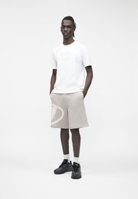 Aukštas žmogus stovėdamas kišenėse rankomis dėvi baltą marškinėlį su iškiliais logotipu, smėlio spalvos šortus su logotipu, baltas kojines ir juodus sportinius batelius.