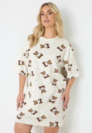 Vrouw draagt een losse, crème kleurige korte mouw jurk bedrukt met meerdere kleine teddyberen en lichtblauwe hartjes.