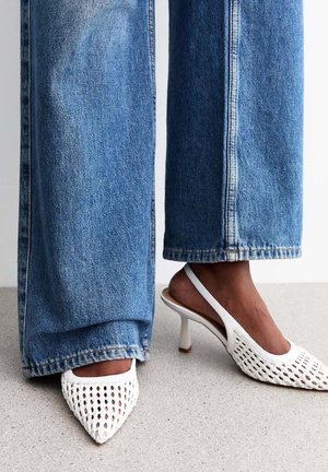 Escarpins blancs à talons avec bride arrière ouverte, bout pointu et design perforé. Portés avec un jean bleu ample.