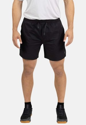 Hombre que lleva pantalones cortos deportivos negros con cordón, calcetines negros y zapatillas negras, de pie contra un fondo claro y liso.