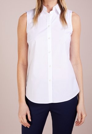 Mouwloze witte blouse met knopen, gedragen met donkere broek, gefotografeerd van schouders tot heupen tegen een neutrale achtergrond.