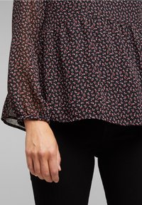Blusa nera a fiori con piccoli fiori rossi e bianchi, maniche lunghe trasparenti, vita arricciata e tessuto morbido e textured.