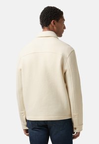 Cremefarvet uldjakke med krave, knapdetaljer og diagonal syning på ærmerne, parret med mørke denimjeans. Simpelt, struktureret design.