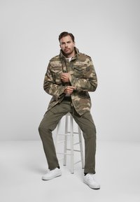 Maskovacia bunda s viacerými vreckami, vysokým golierom a zipsovým zapínaním, kombinovaná s olivovozelenými cargo nohavicami a bielymi teniskami.