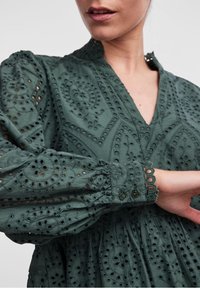 Dunkelgrünes, besticktes Baumwollkleid mit V-Ausschnitt, Ballonärmel und filigranen Lochmustern; hat Knöpfe an den Ärmeln und einen gerafften Rock.