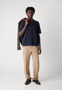 Maglietta navy a righe con colletto a maniche corte, abbinata a pantaloni beige. Giacca in pelle marrone appoggiata sulla spalla sinistra e scarpe scure.