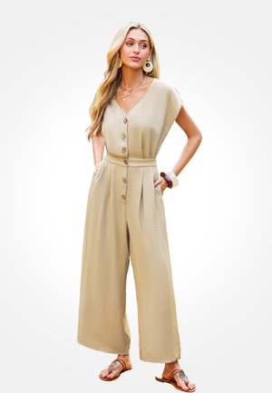 Beige, loszittende jumpsuit met een V-hals, korte mouwen, knopen aan de voorkant, zijzakken en brede pijpen, gecombineerd met bruine sandalen.
