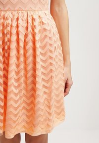 Robe couleur pêche avec une superposition de maille en zigzag, taille froncée, et un bras de femme reposant à ses côtés, sur un fond uni.