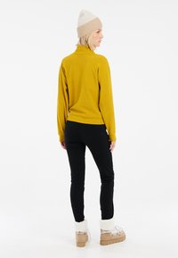 Gelber gerippter Pullover mit hohem Kragen, kombiniert mit schwarzen eng anliegenden Hosen und beigen Stiefeln mit einem flauschigen weißen Bund.