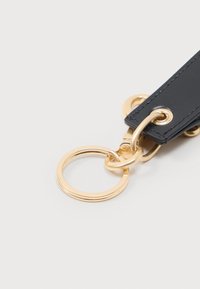 Κοντινό πλάνο χρυσού μπρελόκ προσδεδεμένο σε μαύρο δερμάτινο λουρί με χρυσές γωνιότρυπες σε ανοιχτό φόντο.