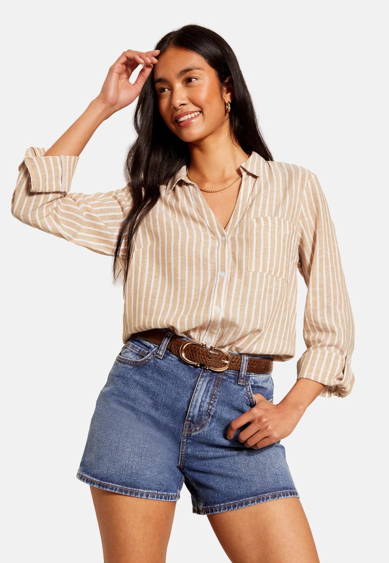 Šviesiai rudos ir baltos spalvos dryžuoti marškiniai su mygtukais ir ilgomis rankovėmis, derinami su aukštakulniais mėlynos džinsinės medžiagos šortais ir rudomis diržu su sagtimi.