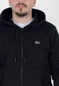 Sudadera con cremallera negra hecha de tela suave, con cordones ajustables, bolsillo canguro y un pequeño logo de cocodrilo bordado en verde.