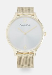 Montre Calvin Klein dorée avec un cadran argenté, design minimaliste, bracelet en mailles et index dorés aux positions 12 et 6 heures.