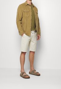 Ljusbrun manchestertröja med två bröstfickor, olivgrön t-shirt, vita shorts med dragsko och grå sandaler med spännen.