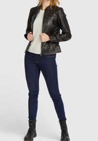 Giacca di pelle nera con design aderente, chiusura con zip e tasche, indossata sopra un top chiaro e abbinata a jeans skinny scuri e stivali neri.