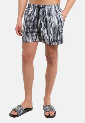 Mand iført sorte og grå mønstrede shorts med snøre og matchende slides på bare fødder, stående med den ene hånd i lommen.