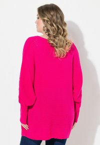 Fuchsia oversized Strickpullover mit geripptem Muster, langen Ärmeln und lockerer Passform, kombiniert mit dunkelblauen Jeans.