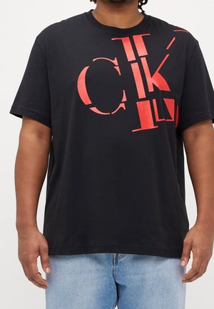Mann trägt ein schwarzes T-Shirt mit großen roten CK-Logo-Buchstaben und hellblauen Jeans, gezeigt vom Hals bis zur Taille.