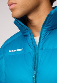 Veste isolante teal avec col haut, fermeture zippée à l'avant et logo "MAMMUT" blanc sur la poitrine. Texture de tissu lisse et brillante.
