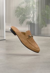 Mocassin en cuir tressé marron clair à enfiler avec détail de boucle en métal, présenté flottant au-dessus d'un sol en béton avec un arrière-plan de stores verticaux flous et de plantes.