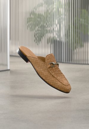 Tannervärvi nööritud nahast kergelt jalga libisev loafer metallist pandla detailiga, kujutatud hõljuvana betoonpõranda kohal, taustal udus vertikaalsed kardinad ja taim.