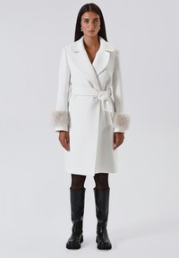 Cappotto bianco con cintura in vita, con morbidi polsini in pelliccia beige, silhouette dritta e revers. Abbinato a stivali neri sopra il ginocchio.
