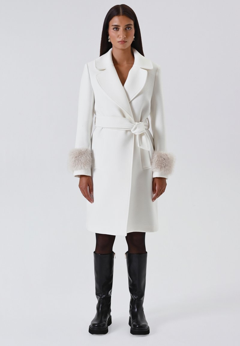 Cappotto bianco con cintura in vita, con morbidi polsini in pelliccia beige, silhouette dritta e revers. Abbinato a stivali neri sopra il ginocchio.
