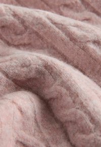 Różowa dzianina, miękka w dotyku, z wzorem w warkocze. Materiał wydaje się puszysty i jest drapowany, ukazując subtelne różnice w kolorze.