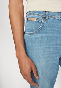 Svijetloplave traperice s glatkom teksturom, s mesinganim detaljima i smeškim kožnim ušivkom s logotipom "Wrangler" na zadnjem džepu.