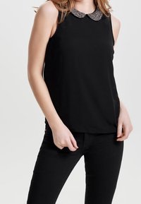 Top nero senza maniche con colletto arrotondato e decorazione metallica. Tessuto morbido; taglio dritto, indossato con pantaloni neri.