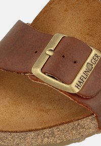 Sandal in pelle marrone con una larga cinghia e una fibbia in ottone. La soletta è in sughero texturizzato con un rivestimento liscio di colore neutro.
