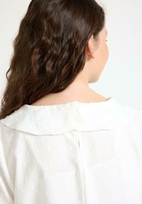 Person med långt, vågigt brunt hår som bär en vit blus med struktur och bred krage, sedd bakifrån mot en neutral bakgrund.
