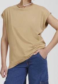 Béžové oversize tričko s krátkými ohrnutými rukávy, vyrobené z měkké látky; nošeno s modrými džínovými kalhotami. Jednoduchý design, žádné viditelné vzory.