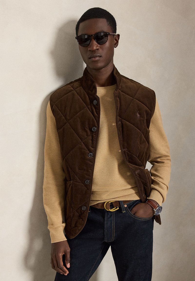 Uomo con occhiali da sole scuri, maglione color sabbia, gilet trapuntato marrone, jeans scuri con cintura, in piedi contro un muro chiaro e uniforme.