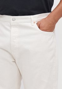 Pantalones de algodón blancos con pierna recta, que cuentan con un bolsillo frontal, acentos de botón de cobre y una textura sutil.